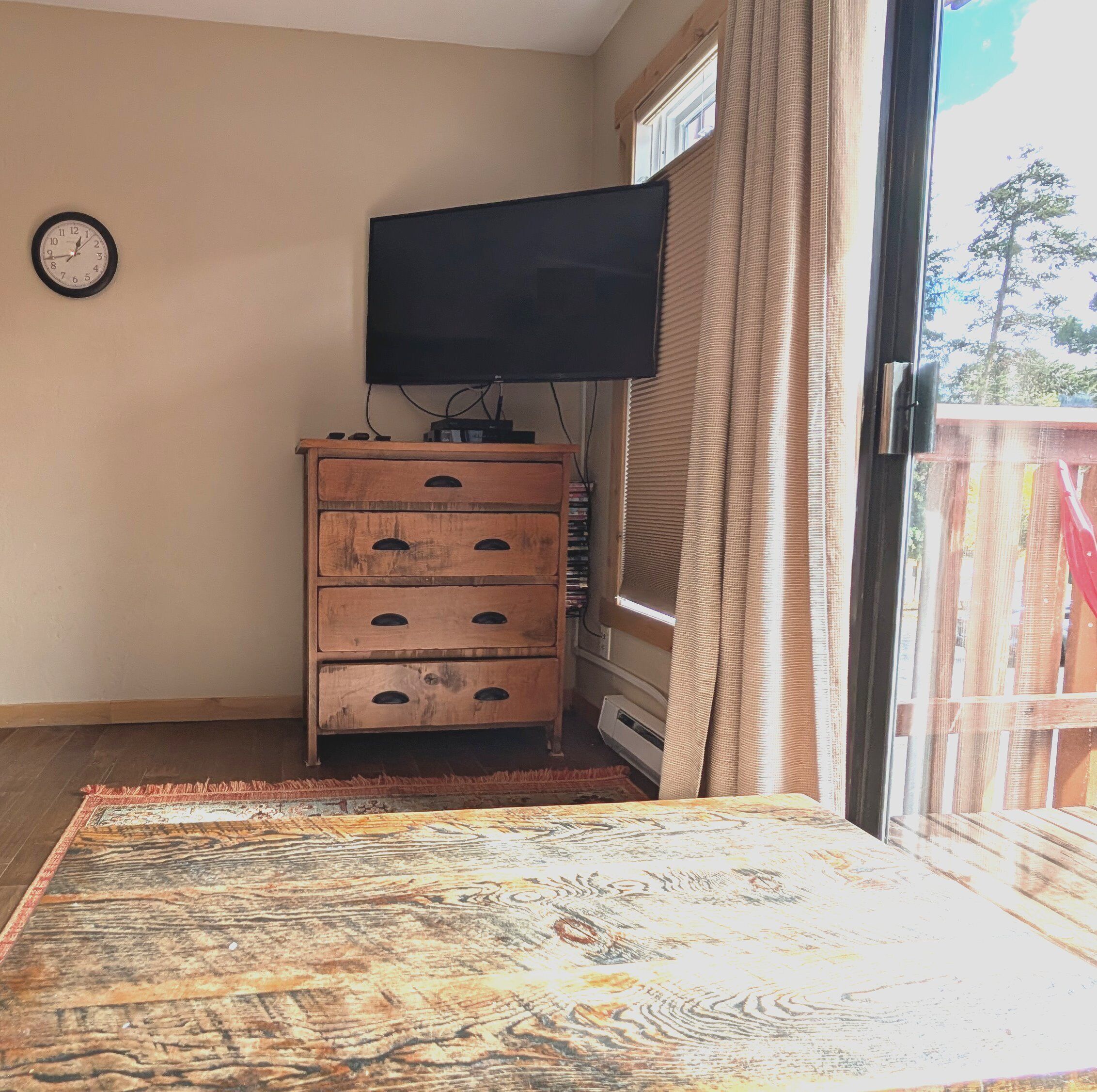 Una habitación con un aparador de madera, un televisor de pantalla plana montado en la pared, una alfombra estampada y un ventanal corredero que da a un espacio exterior.