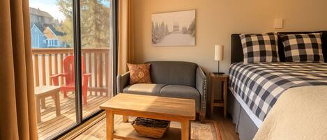 A room with a bed, a sofa, a table, and a window overlooking a deck.