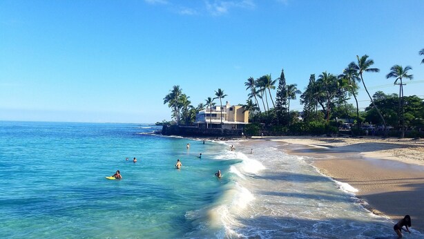A beach with clear blue water, people swimming and surfing, and a building with a balcony overlooking the ocean.
