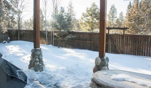 Une pergola en bois, avec des piliers en pierre, se dresse dans une cour arrière enneigée, bordée d’une clôture en bois et avec des arbres en arrière-plan.