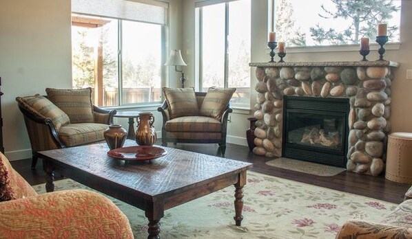 Un salon chaleureux avec une cheminée en pierre, deux fauteuils, une table basse et de grandes fenêtres.