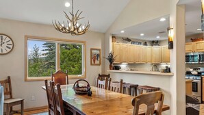 A dining area with a wooden table and chairs, a chandelier, and a view of greenery outside.