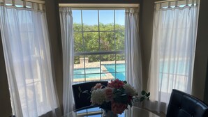 Une pièce avec une table en verre, un vase rempli de fleurs et une vue sur une piscine, visible à travers une fenêtre ornée de rideaux légers.