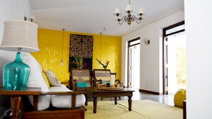 Ein Zimmer mit gelber Akzentwand, einem Holztisch, einem Sofa mit weißen Kissen, einer türkisblauen Vase und einer Kristallleuchter.