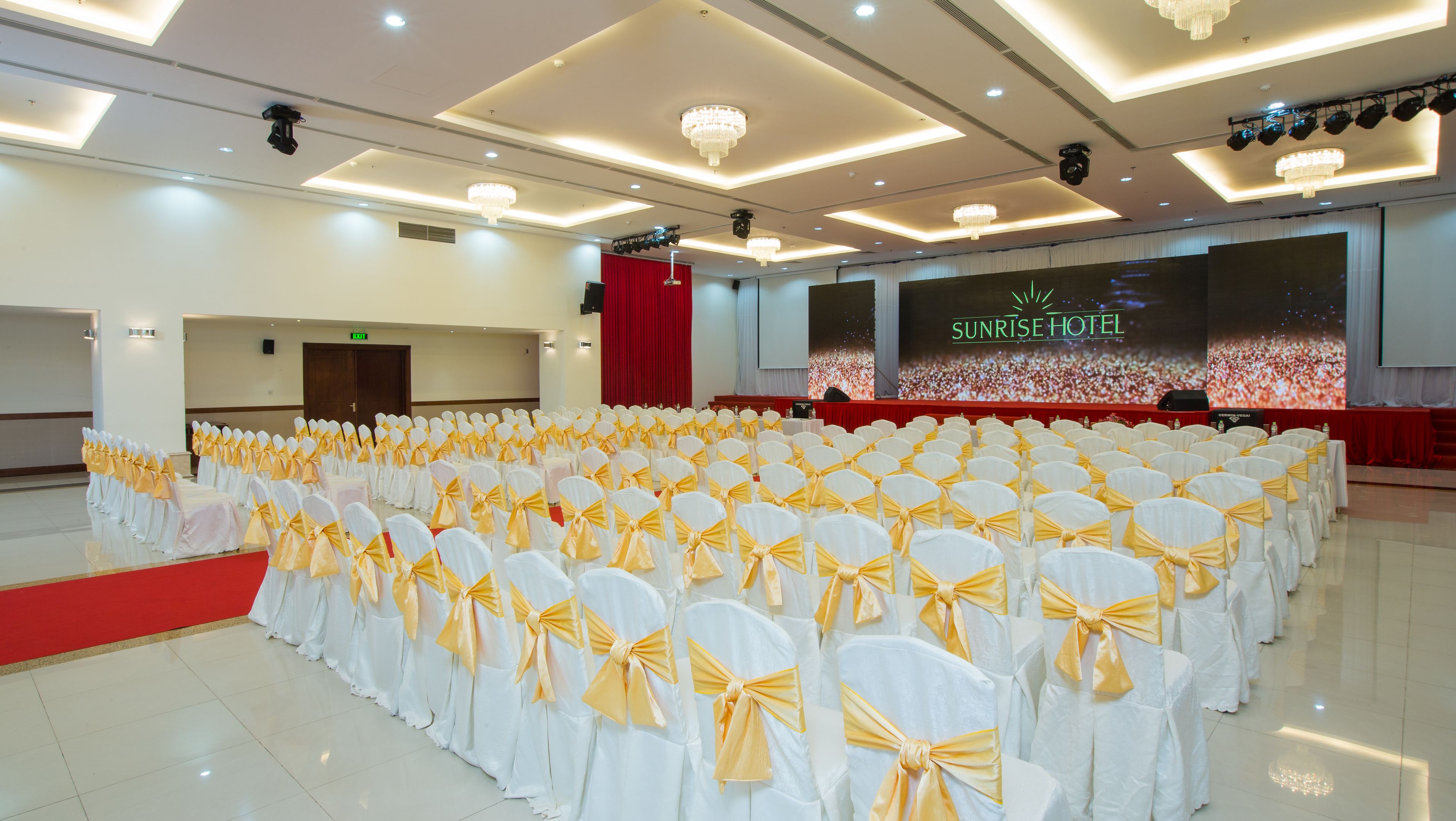 A large hall with rows of white chairs tied with yellow sashes, a red carpet, and a stage with a large screen displaying the Sunrise Hotel logo.