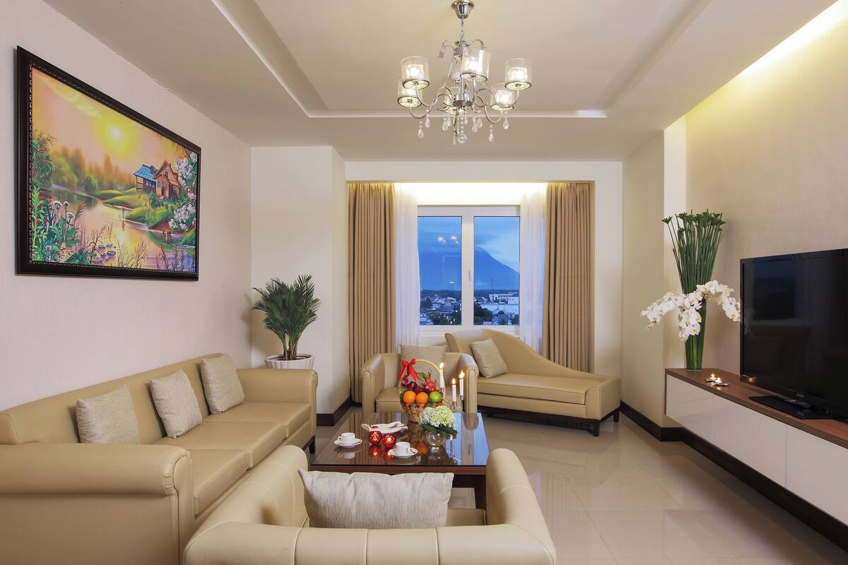 A living room with a sofa, a coffee table with fruits and candles, a TV, a painting, and a view of a mountain through a window.