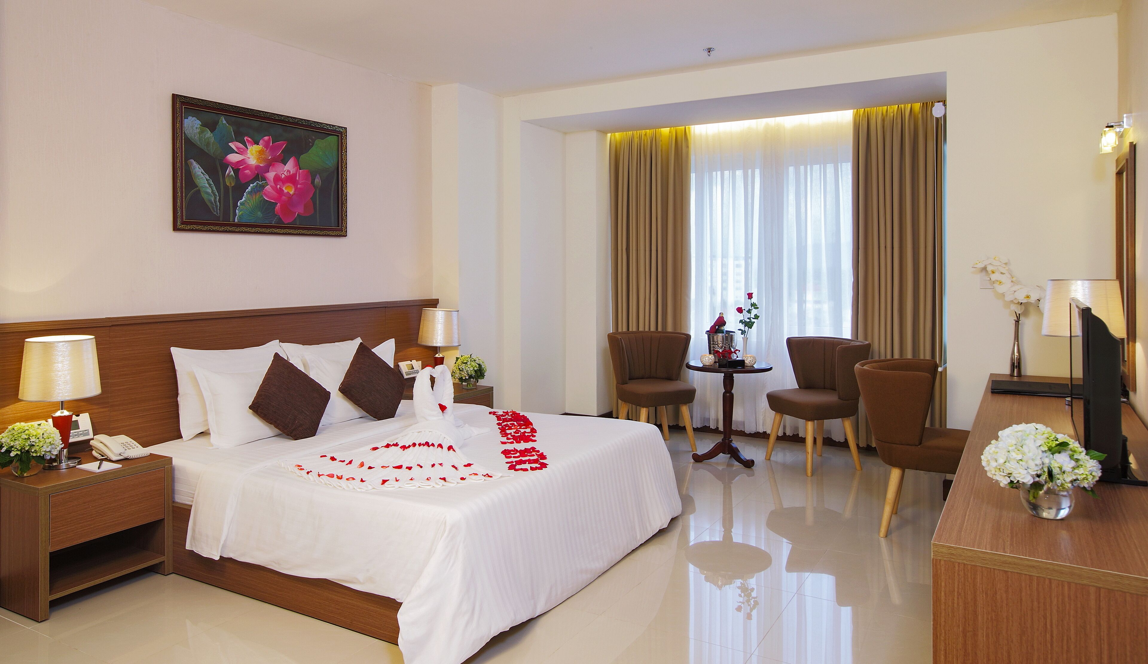 A hotel room with a neatly made bed, a small table with a vase of flowers, two chairs, a desk with a lamp, and a painting on the wall.