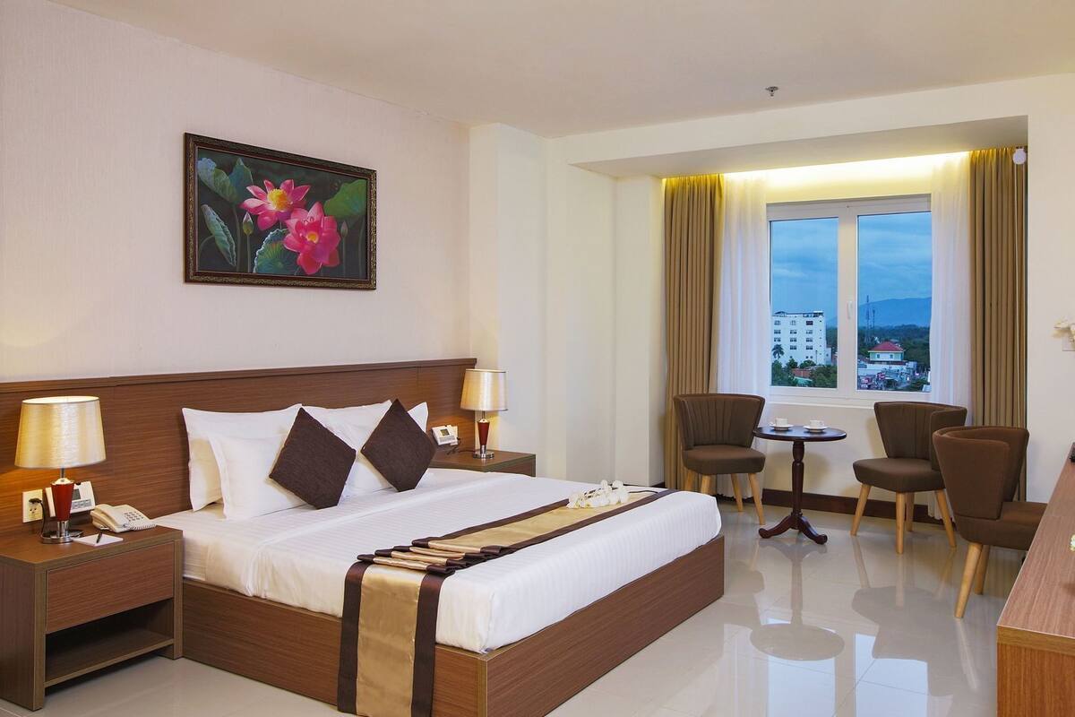 A hotel room with a bed, bedside tables, a desk, a chair, a small table, and a window with curtains.