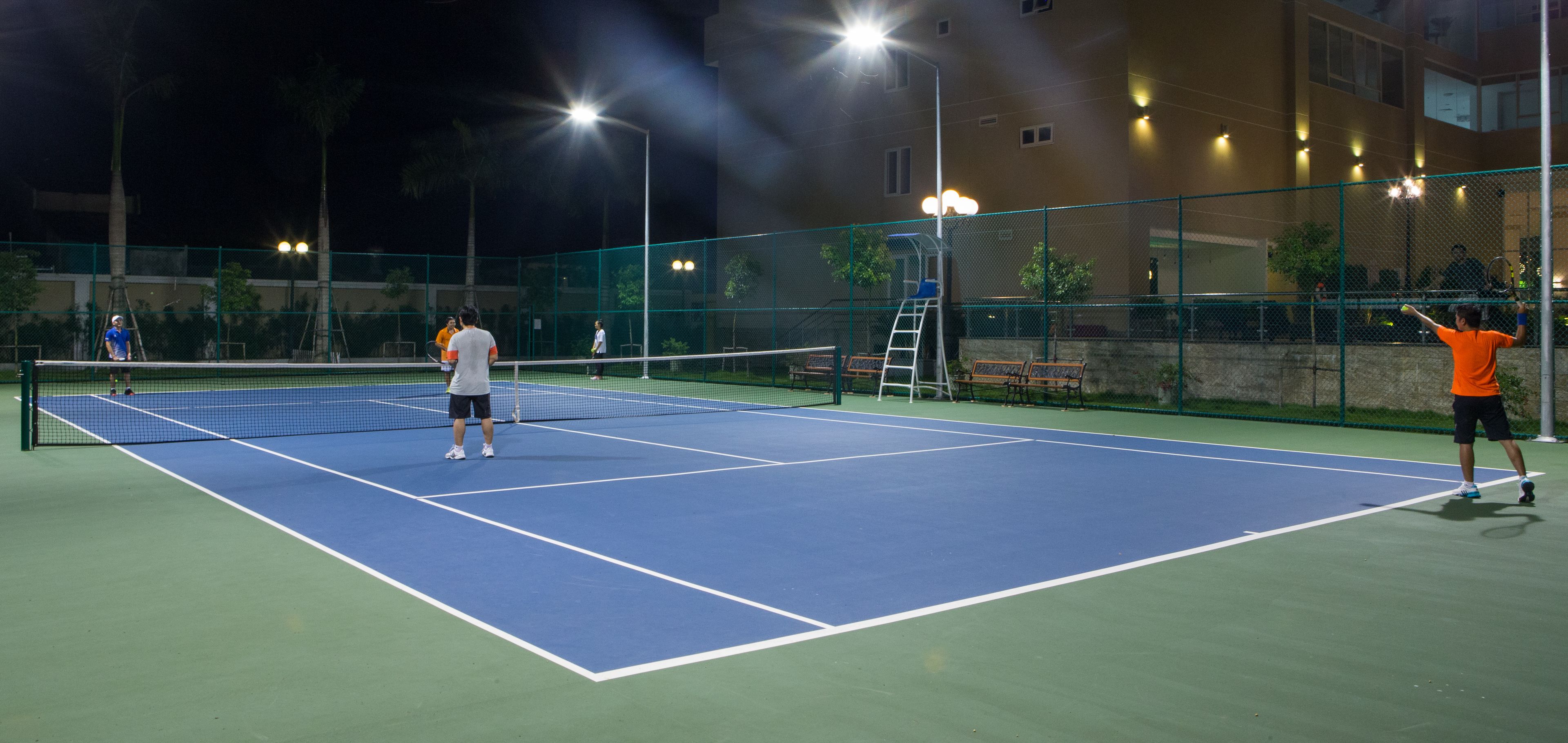 An outdoor tennis court at night with a net, two players, and a ladder.