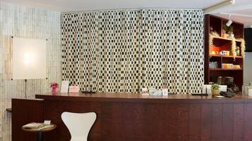 A reception area with a patterned wall, a wooden reception desk, a white chair, and a shelf with decorative items.