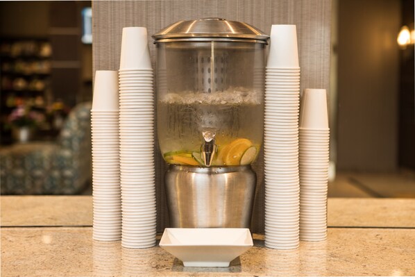 A beverage dispenser with a metal container, surrounded by stacks of white paper cups. - West Wing at Park Town (Saskatoon)