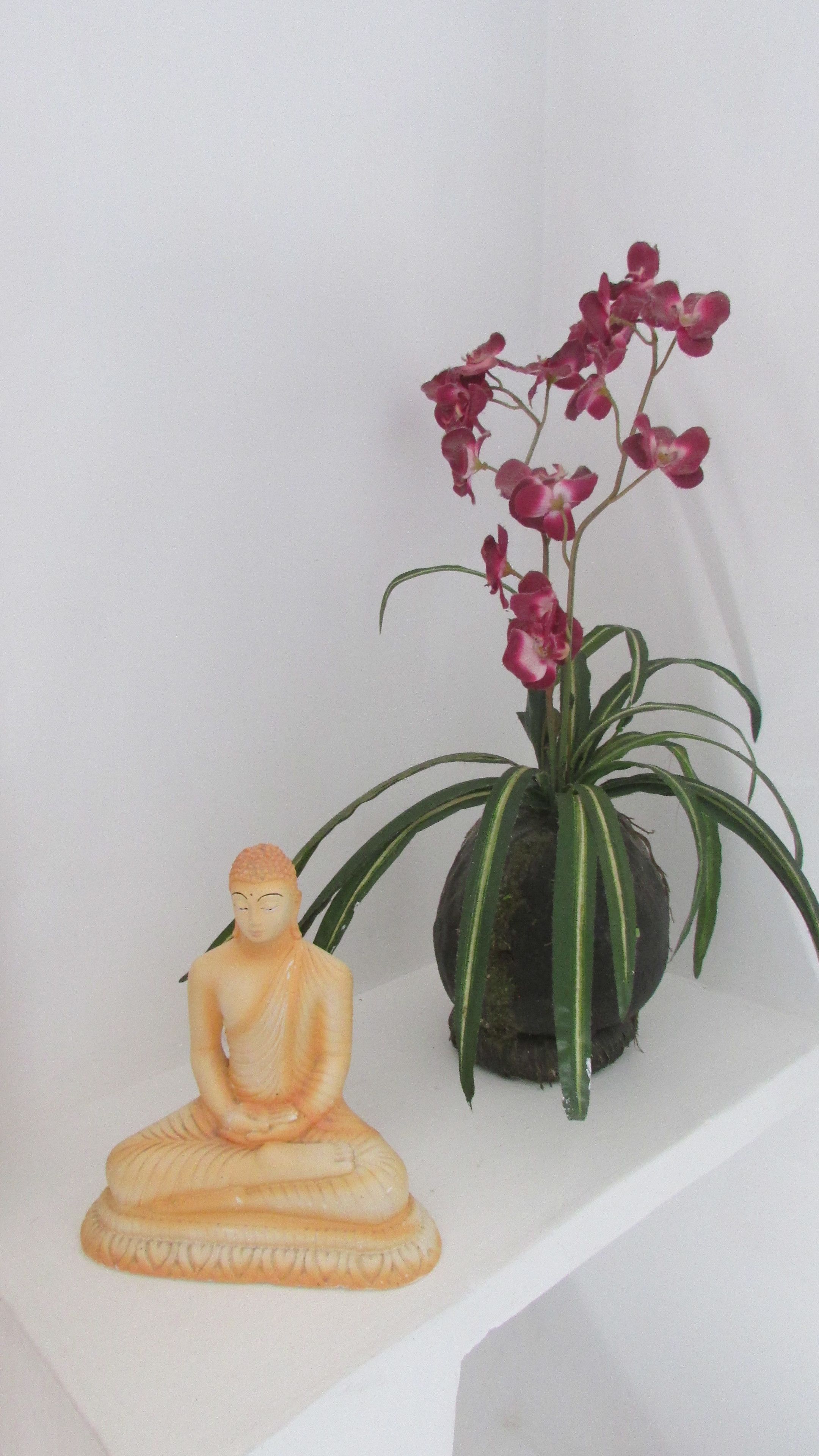 Una estatua de Buda y una orquídea en maceta sobre un estante blanco.