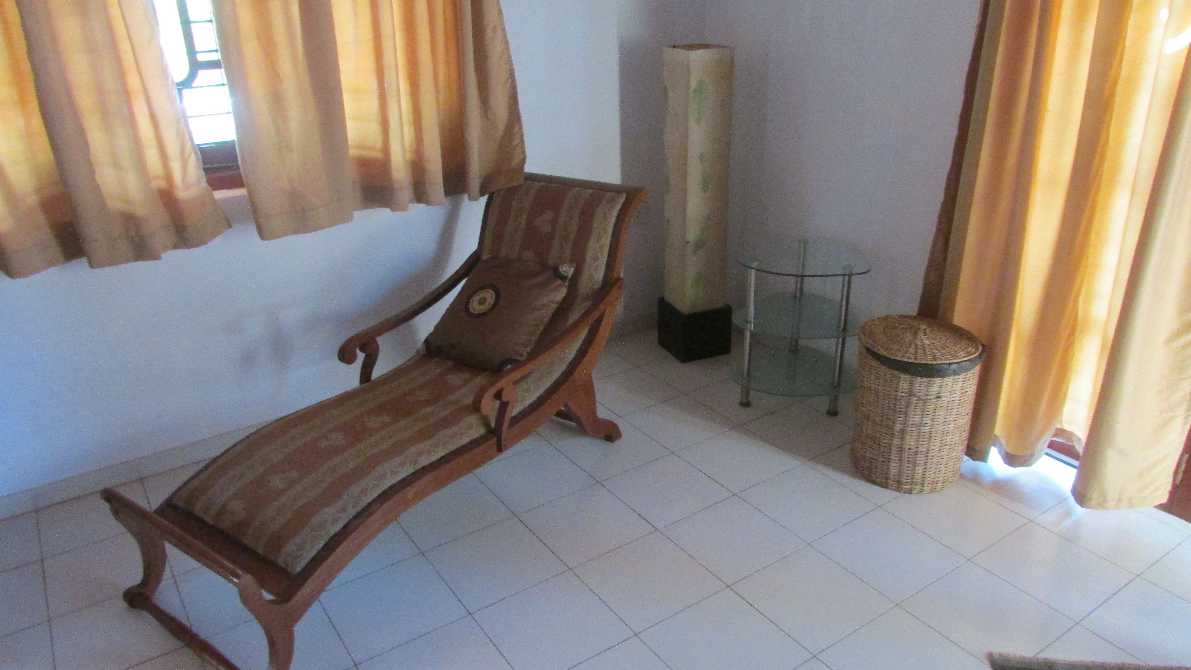 A sun lounger, a glass table, a wicker basket, and a tall, narrow cabinet in a room with tiled flooring and curtains.