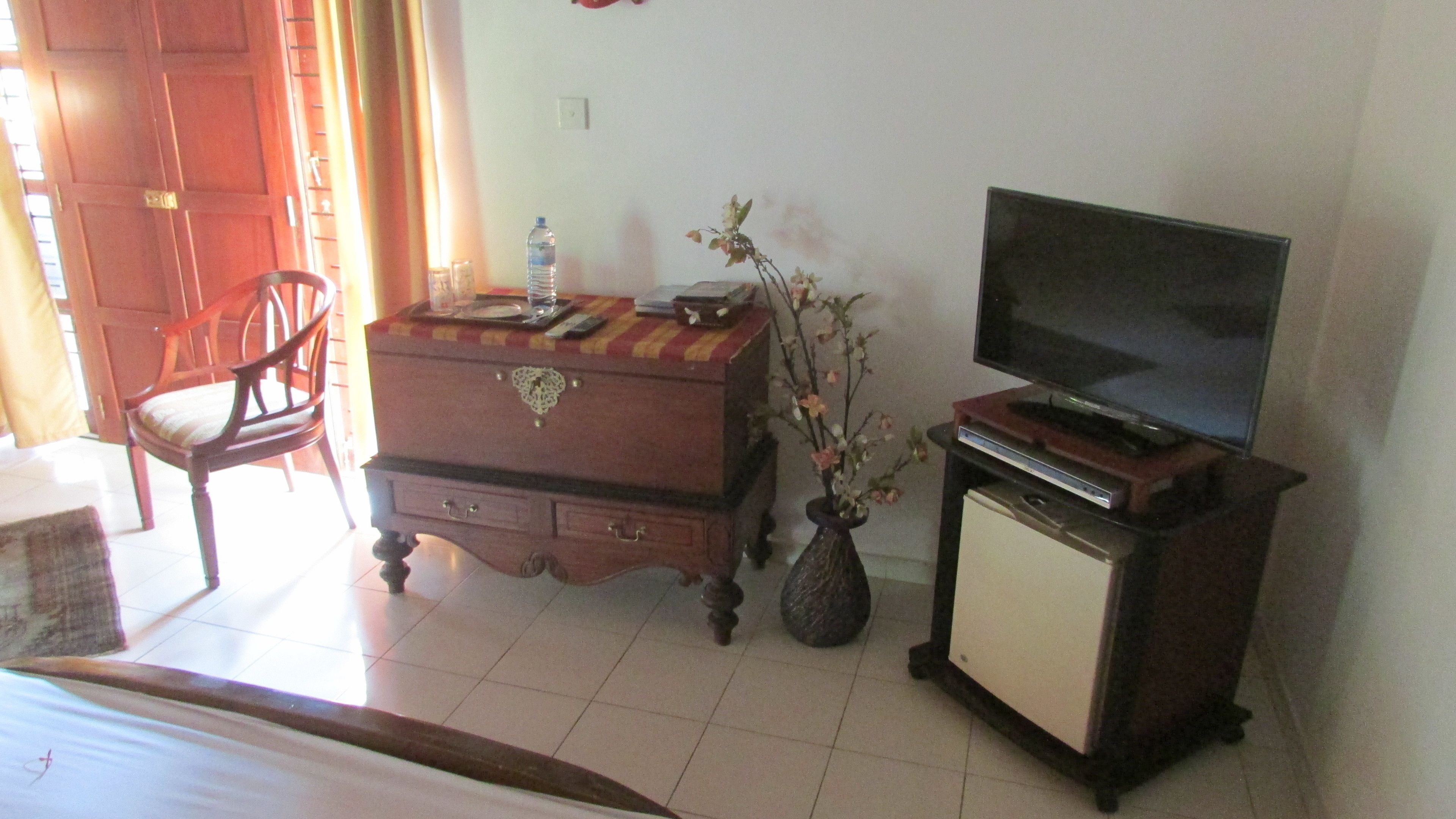 A room with a wooden dresser, a television on a stand, a chair, and a vase with flowers.