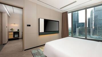 A modern hotel room with a large bed, a flat-screen TV mounted on the wall, a city view through large windows, and a small kitchenette area.