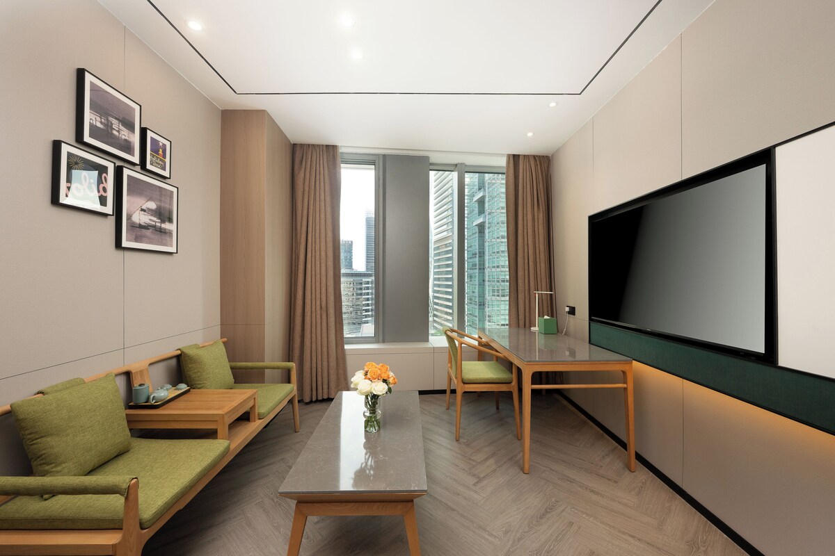 A modern hotel room with a sofa, a dining table, a flat-screen TV, and a large window with a city view.