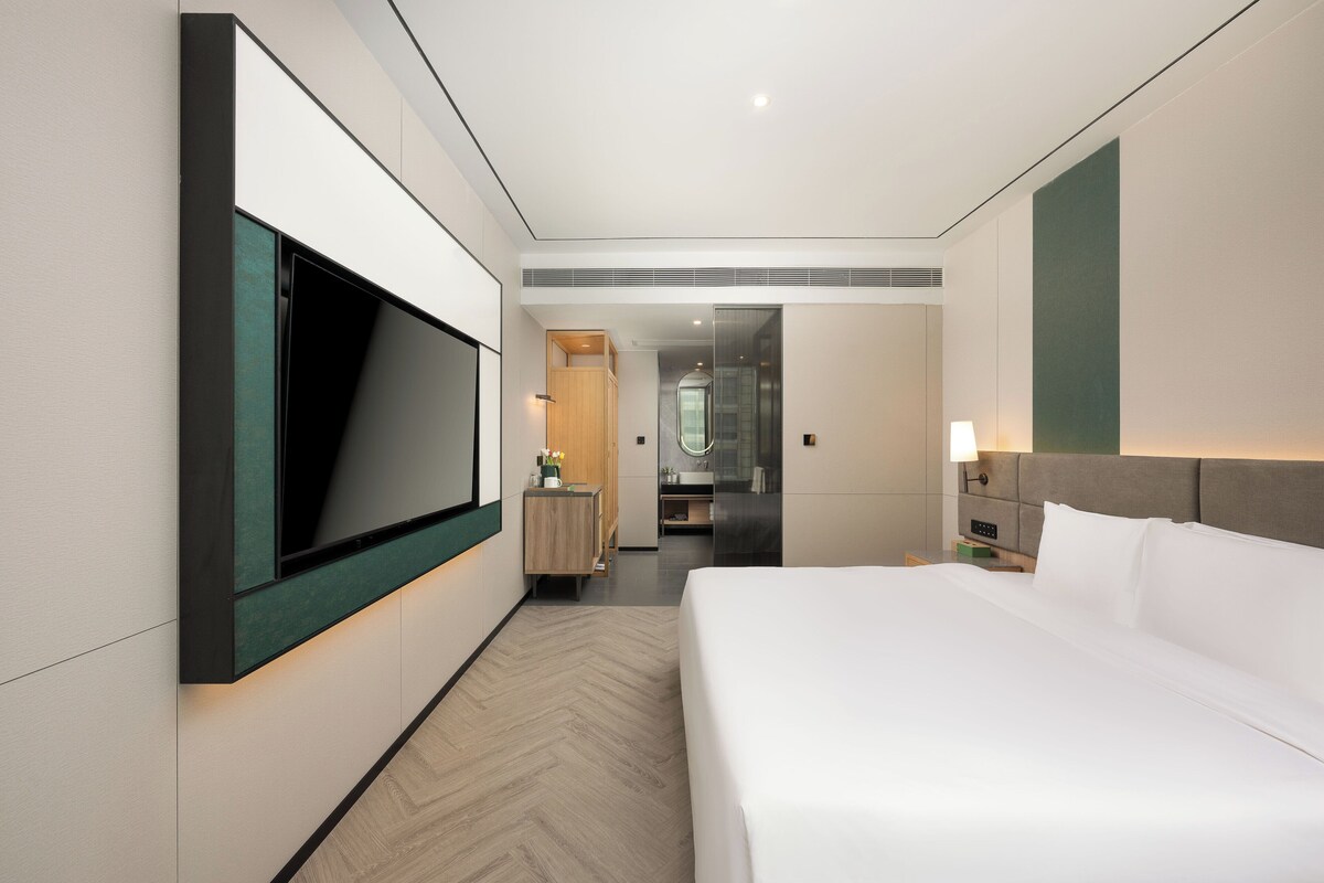 A modern hotel room with a large bed, a TV mounted on the wall, a vanity with a mirror, and a bathroom visible through an open door.