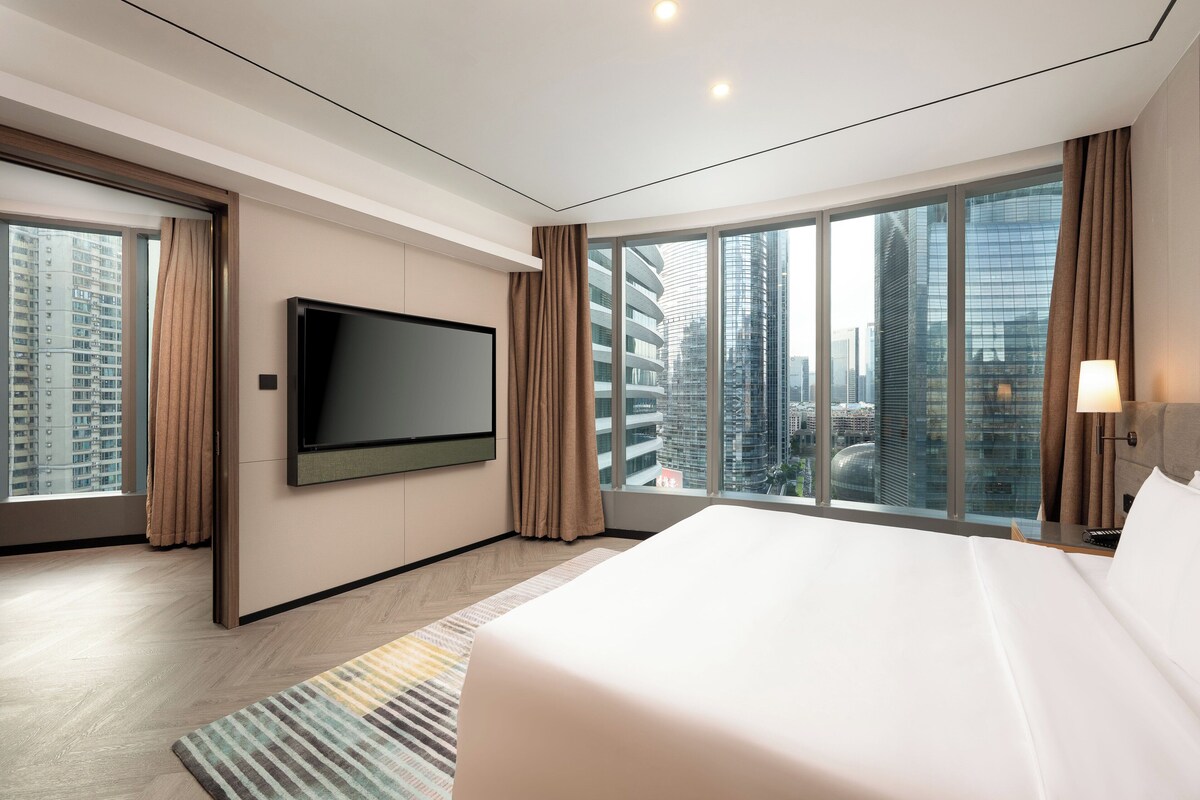 A modern hotel room with a large bed, a flat-screen TV mounted on the wall, and a city view through large windows.