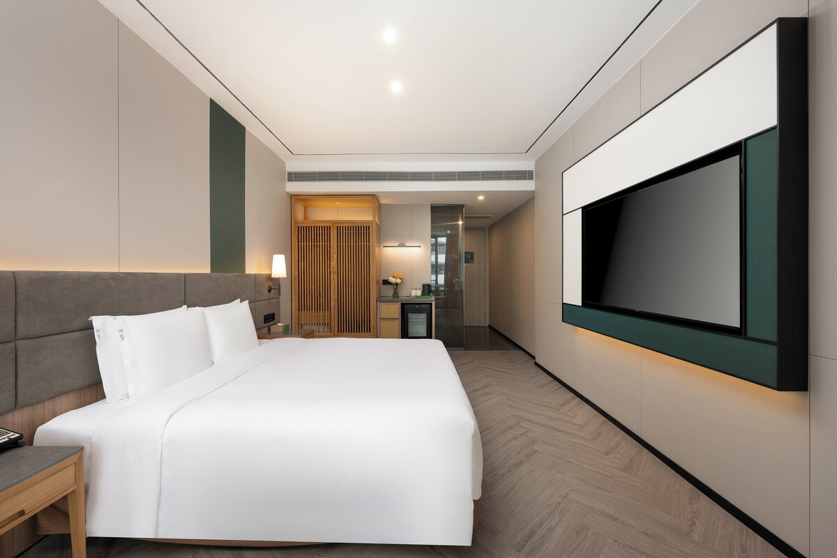 A modern hotel room with a large bed, a TV mounted on the wall, a desk, and a small kitchenette.