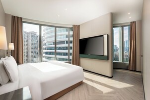 A modern hotel room with a large bed, a flat-screen TV mounted on the wall, and large windows offering a city view.