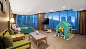 Una habitación de hotel moderna con un ventanal grande que ofrece vistas a la ciudad, un televisor de pantalla plana, una casita de juegos verde y una zona de estar con mesa y sillas.