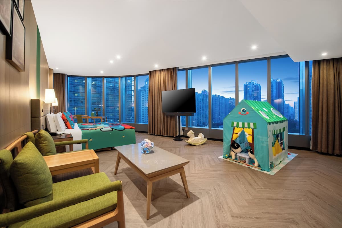 Una habitación de hotel moderna con un ventanal grande que ofrece vistas a la ciudad, un televisor de pantalla plana, una casita de juegos verde y una zona de estar con mesa y sillas.