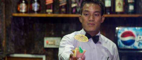 Een bartender houdt een cocktail met een citroen twist en een groene garnering vast, staand voor een bar met drankflessen op de planken.