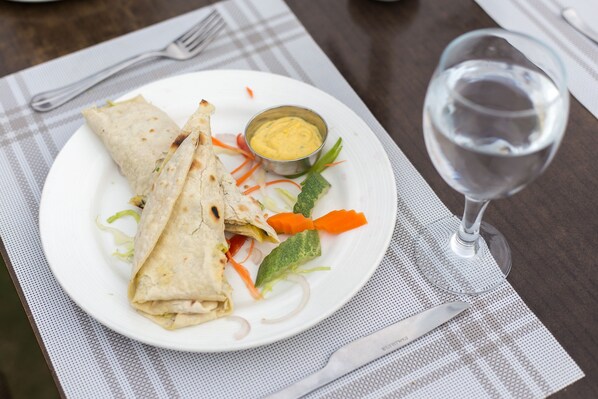 Une assiette contenant un wrap, un petit bol de sauce et un verre d’eau, posée sur une table avec une fourchette et un couteau.