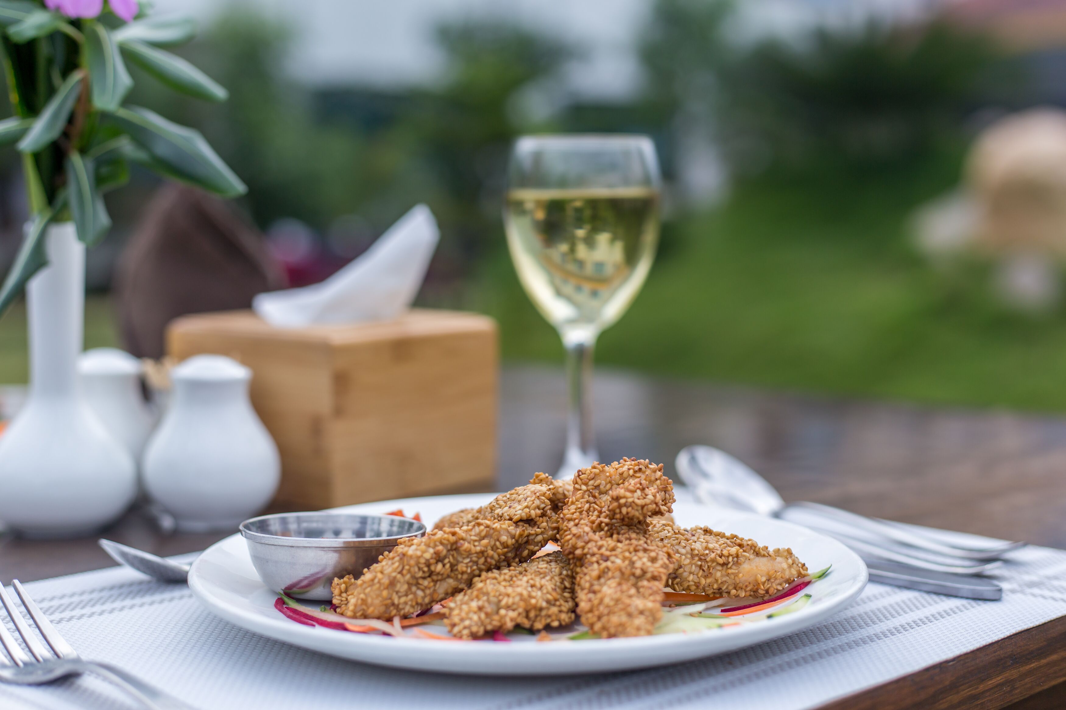 A plate of sesame chicken with a glass of white wine, salt and pepper shakers, and a napkin on a table.