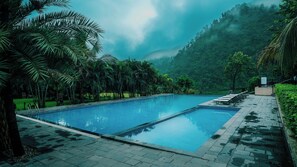 Une piscine rectangulaire dont les bords sont recouverts de carreaux, entourée d’une allée pavée, de palmiers et dominée par une colline boisée enveloppée de brume.