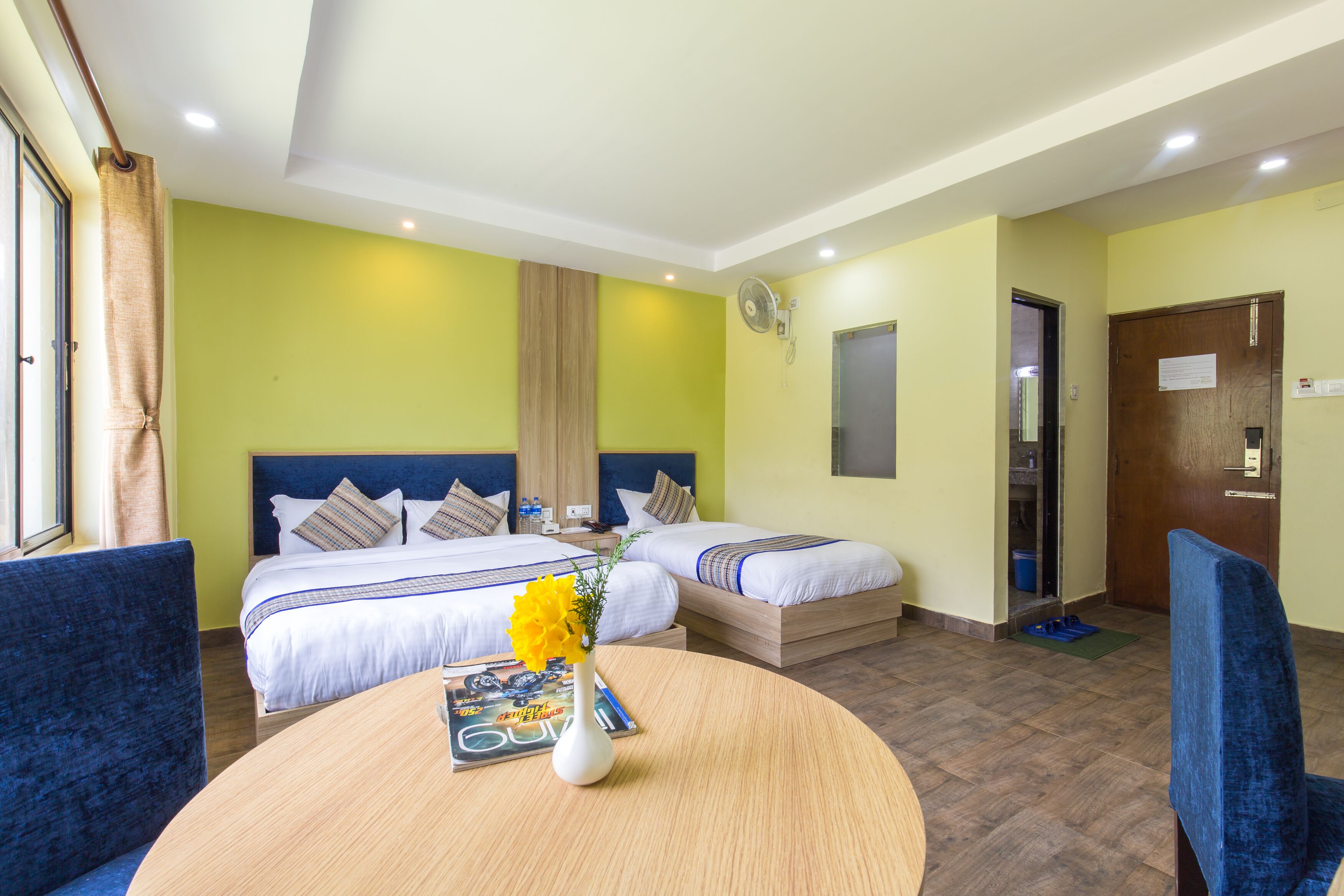 Quarto de hotel com duas camas, uma mesa redonda com um vaso e revista, e uma cadeira azul.