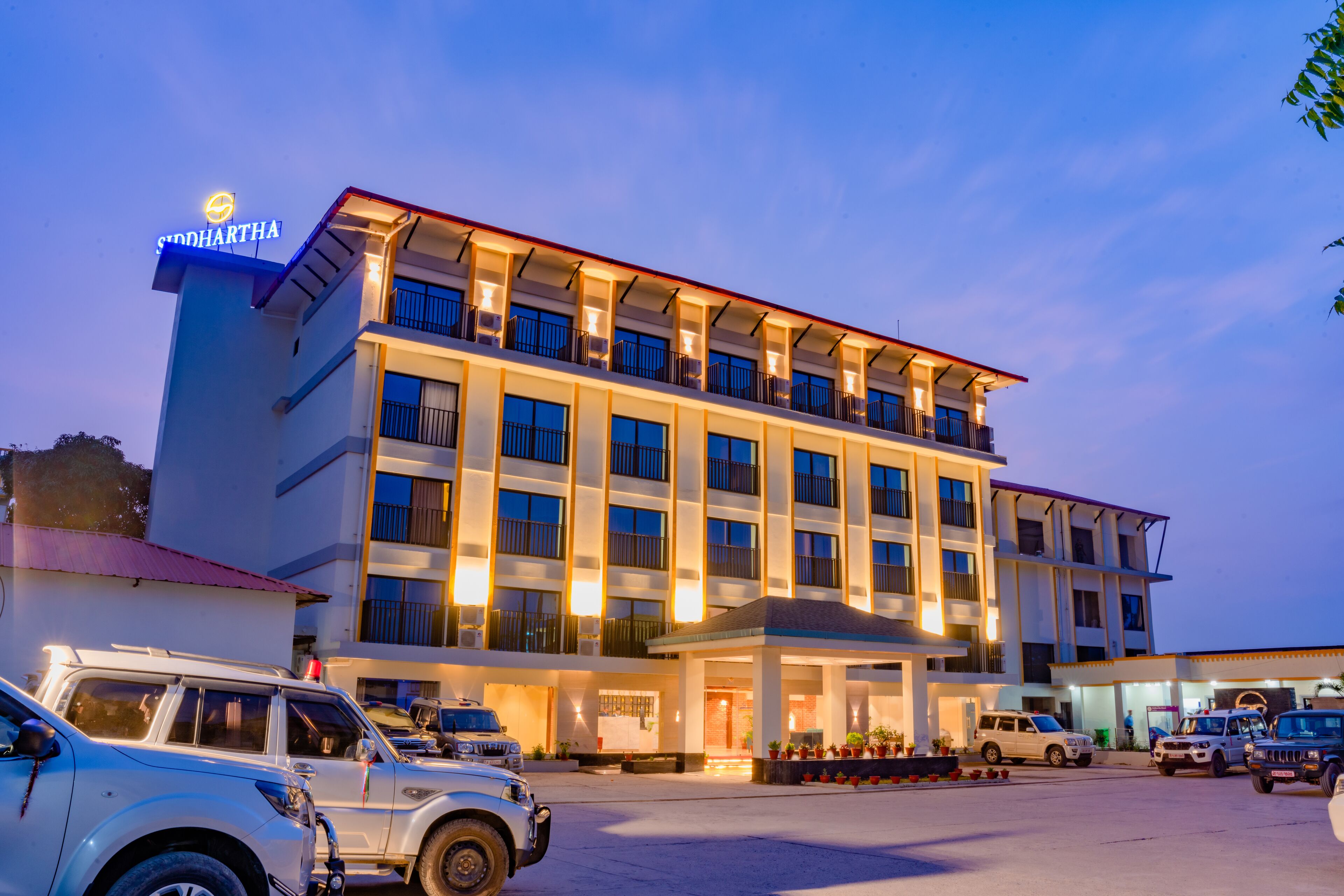 A multi-story hotel with a prominent sign, multiple balconies, and a parking area with several vehicles.