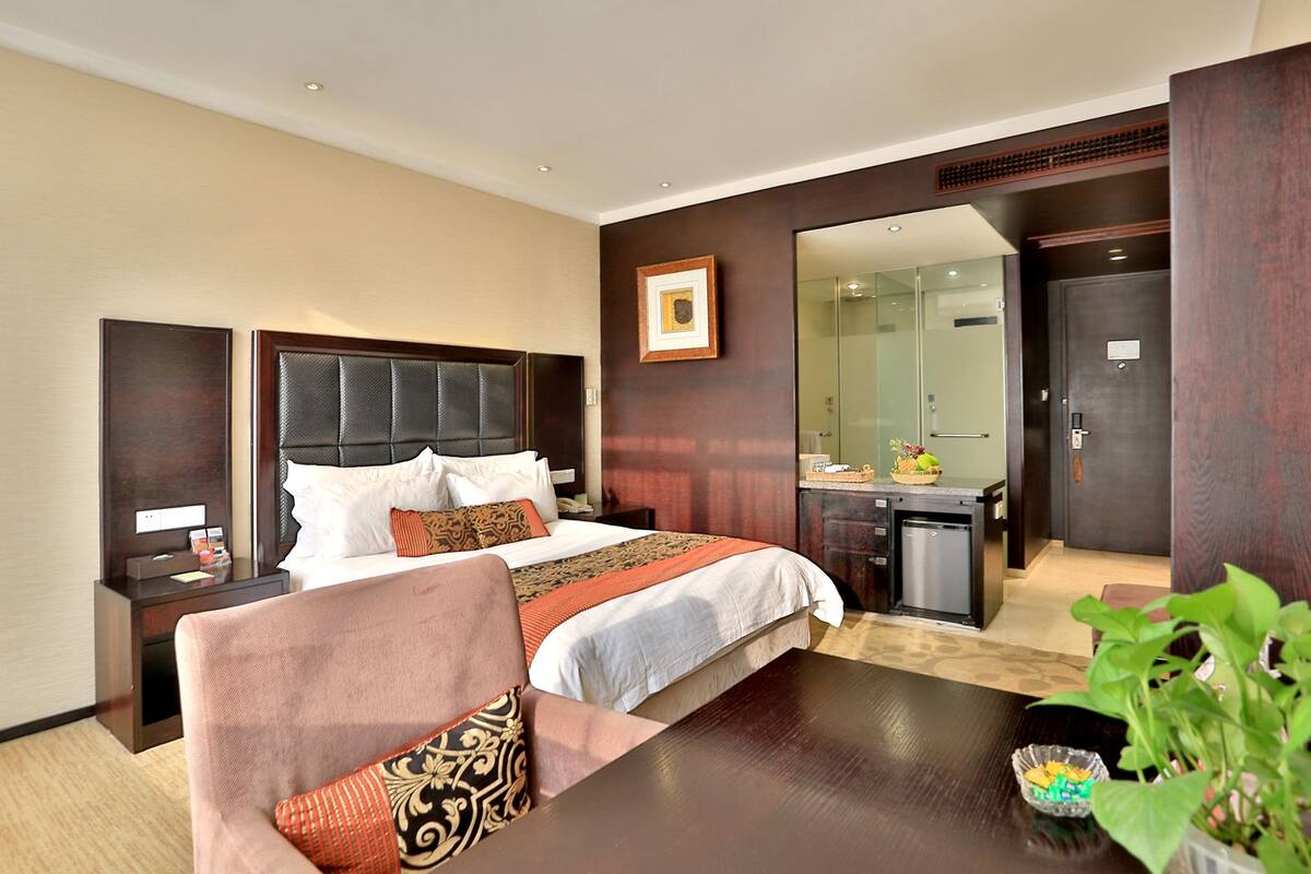 A hotel room with a large bed, a desk, a chair, a small table, and a plant.