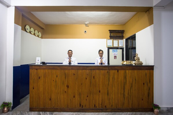 A reception desk with two staff members, a clock, a framed certificate, and a small statue.