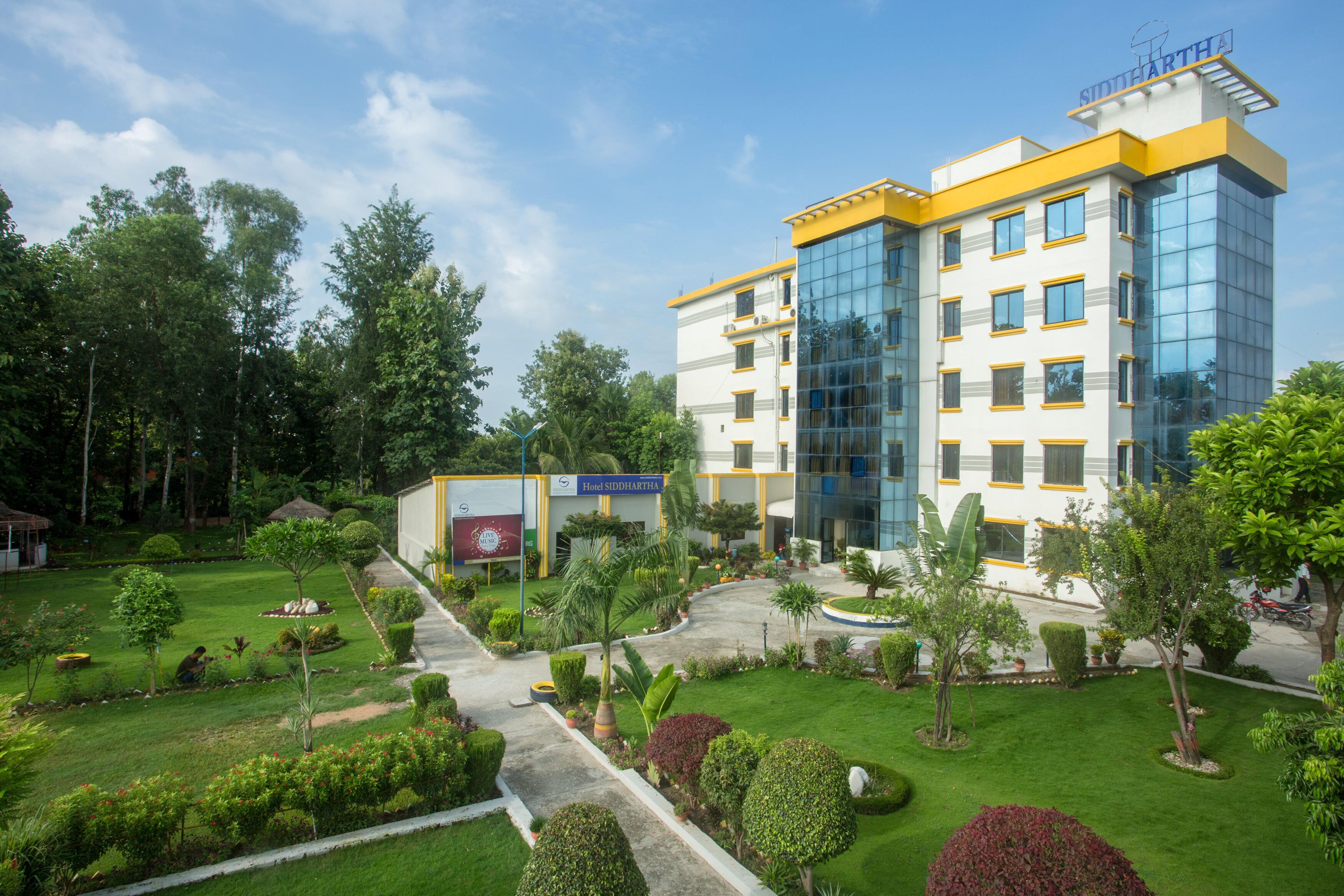 Een hotel met modern design, met een grote glazen gevel en een verzorgde tuin met diverse planten en een pad naar de ingang.
