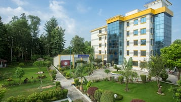 Een hotel met modern design, met een grote glazen gevel en een verzorgde tuin met diverse planten en een pad naar de ingang.