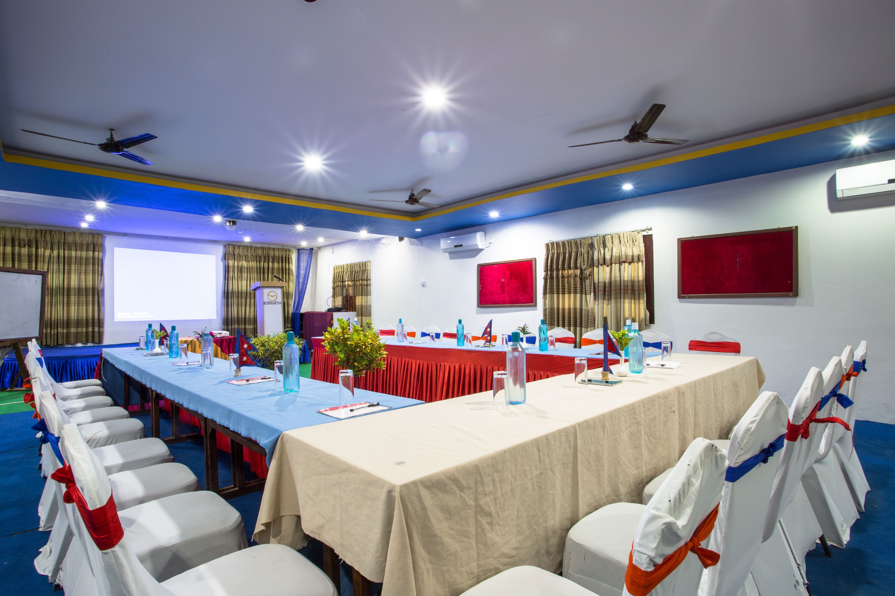 Sala de conferências com uma mesa comprida, cadeiras, ventiladores de teto e uma tela de projetor.