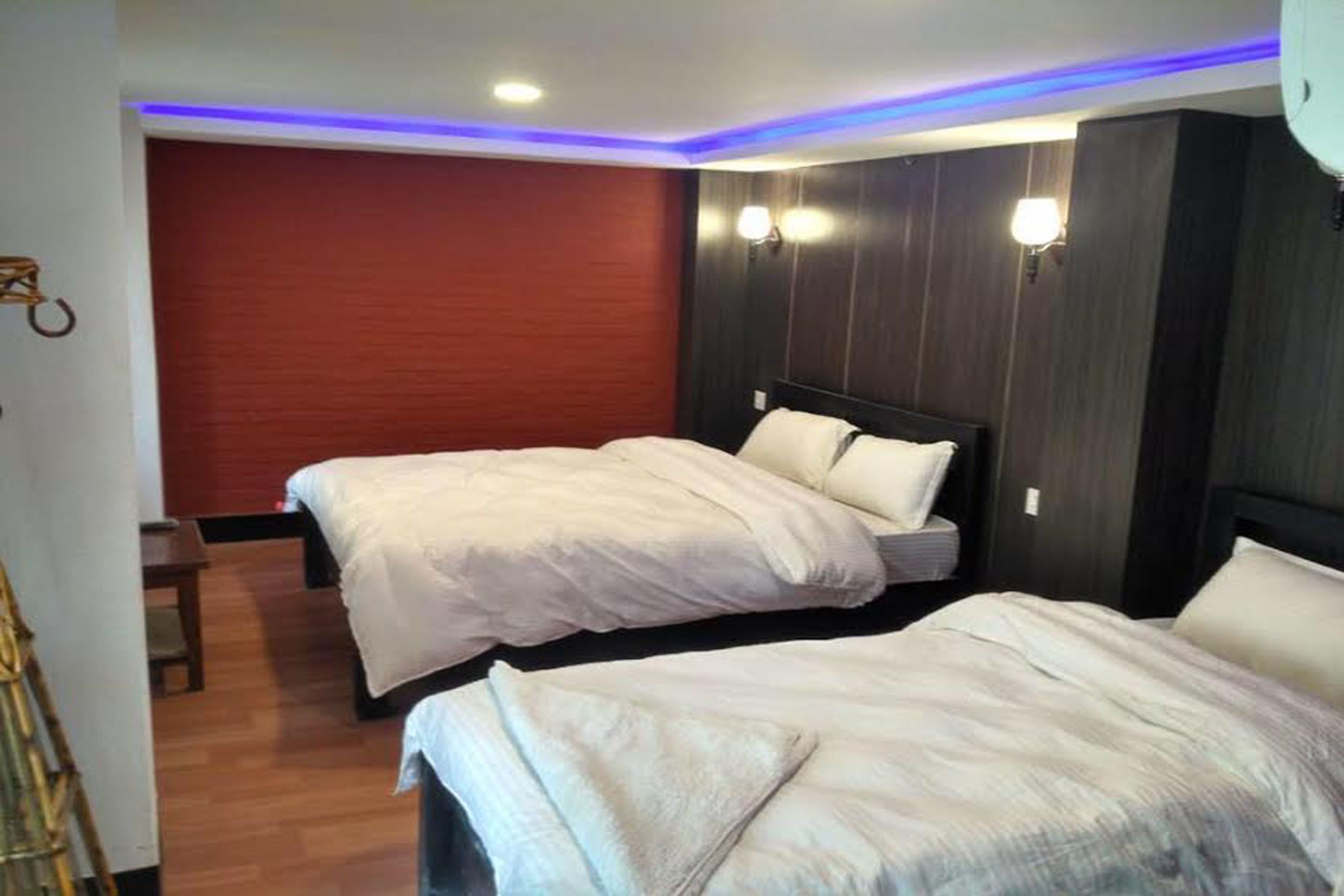 A hotel room with two beds, wooden flooring, a red accent wall, and wall-mounted lighting.