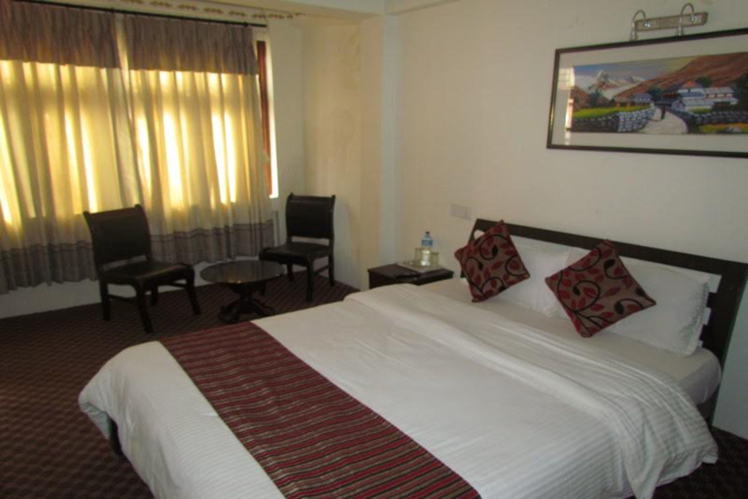 A hotel room with a large bed, a small round table, two chairs, a nightstand with a water bottle, and a painting on the wall.