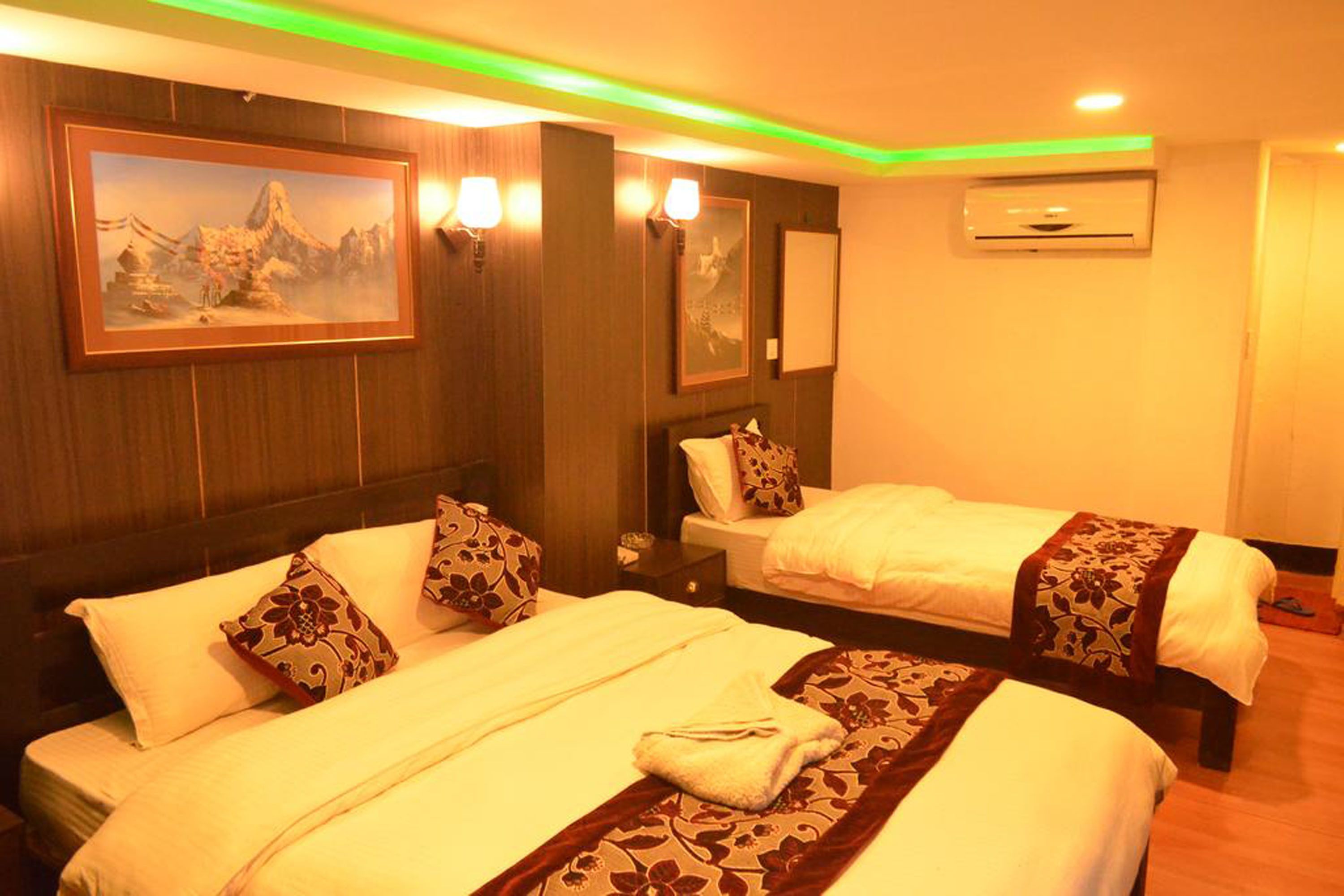 A hotel room with two beds, each with a white bedspread and decorative pillows. There are wall-mounted lamps, a framed painting, and an air conditioning unit.