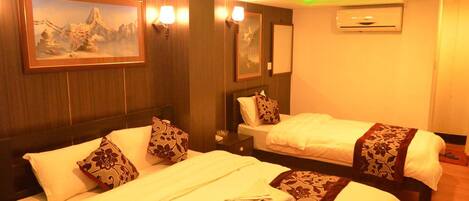 A hotel room with two beds, each with a white bedspread and decorative pillows. There are wall-mounted lamps, a framed painting, and an air conditioning unit.
