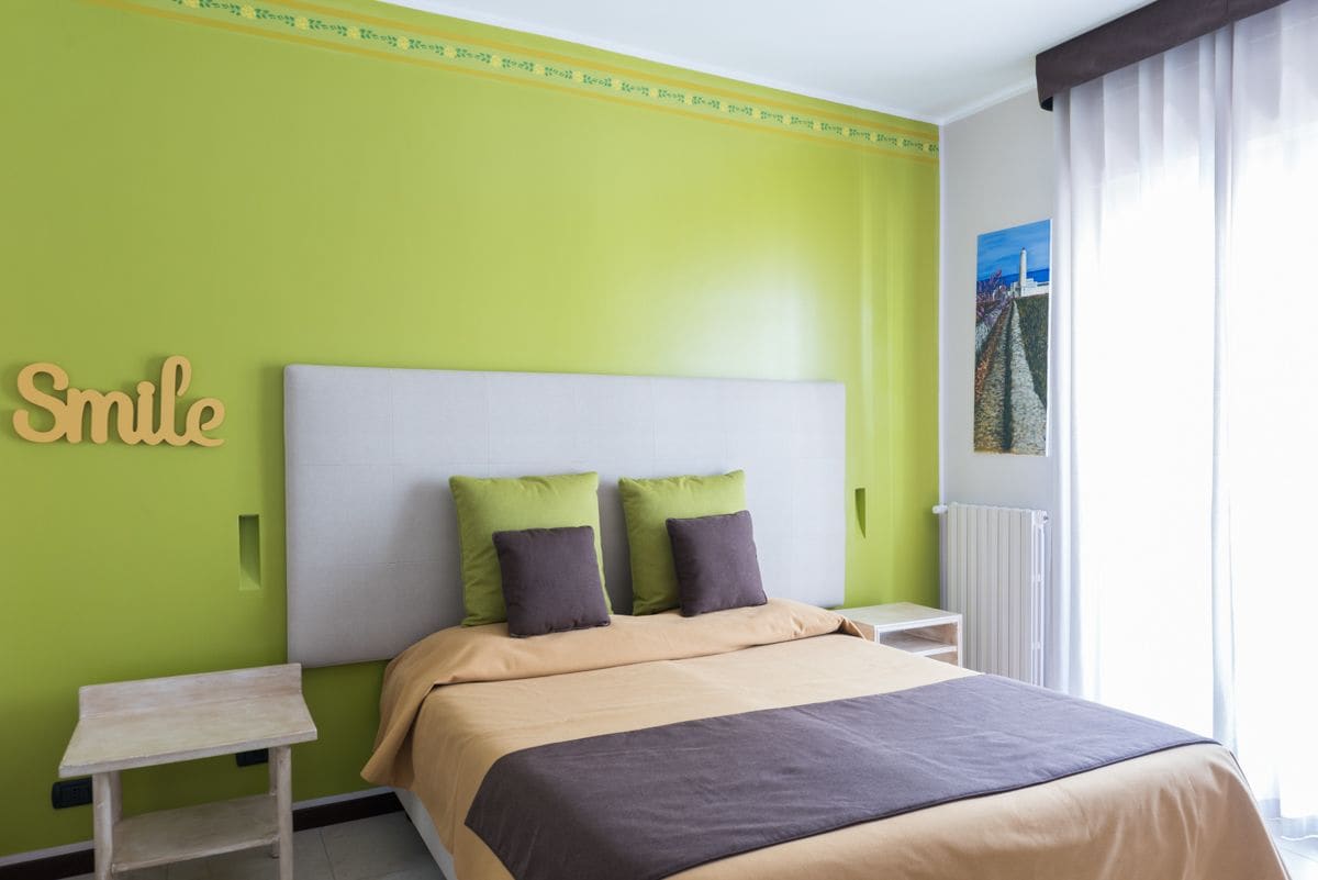 Una camera da letto con una parete verde, un letto con testiera bianca, due cuscini, un comodino e un quadro appeso al muro.