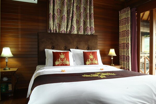 A hotel room with a wooden headboard, two bedside lamps, a bed with white linens and red pillows, and a window with curtains. - Puri Karang Besakih (Rendang)