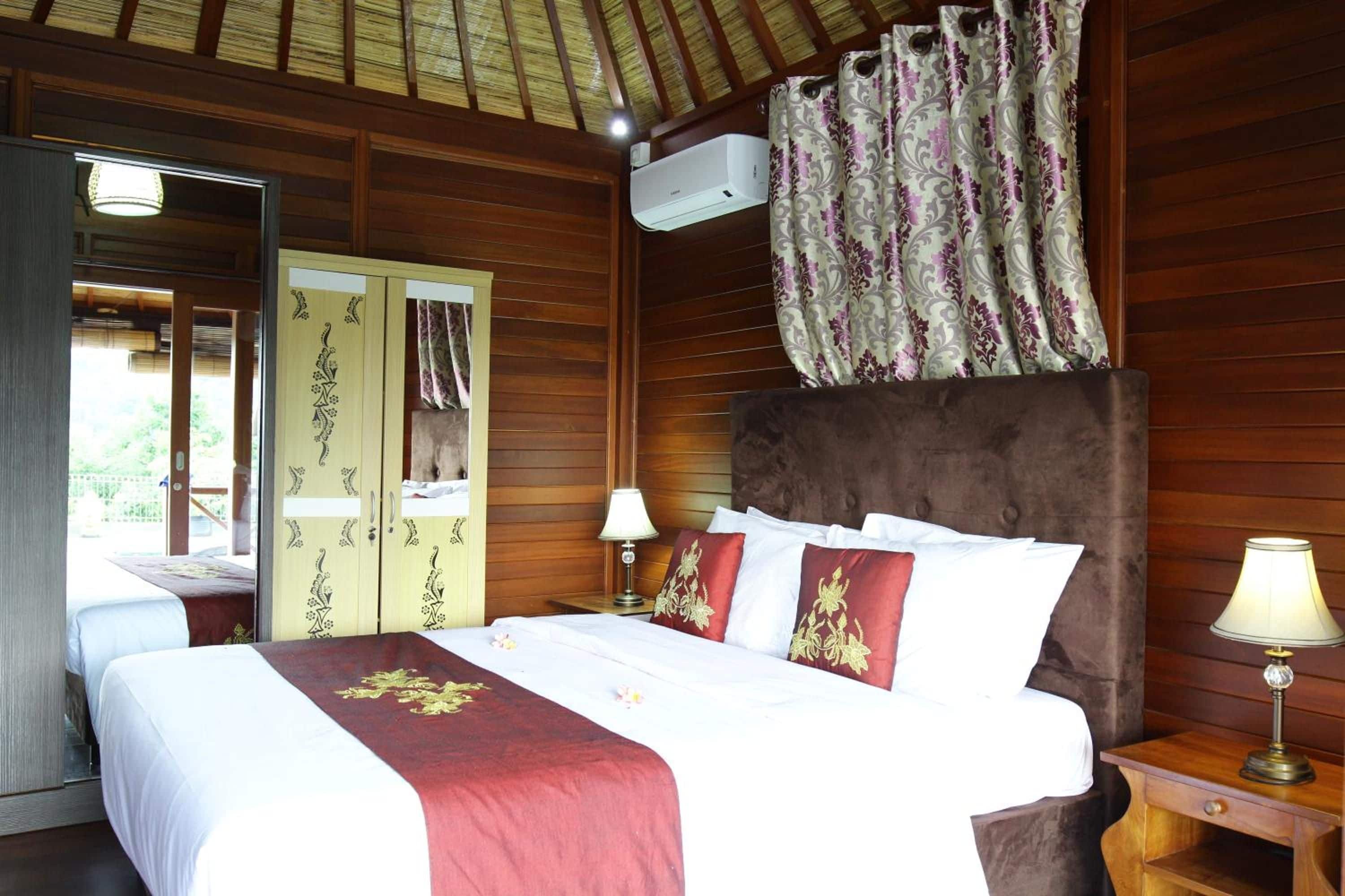 A hotel room with a wooden ceiling, a bed with white bedding and red pillows, a nightstand with a lamp, and a window with curtains.