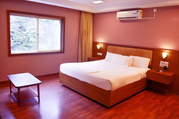 Una cama bien hecha con sábanas blancas, una mesita de noche de madera, una pequeña mesa y una ventana con cortinas.