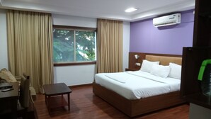 Ein Hotelzimmer mit einem Holzbett, einem kleinen Tisch, einem Sessel, einem Fenster mit Vorhängen und einer Klimaanlage.