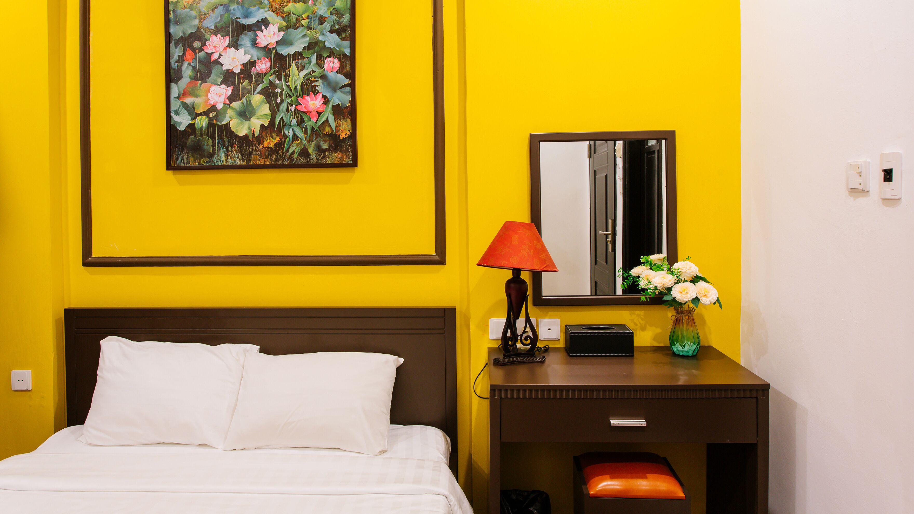 Ein Hotelzimmer mit Bett, Nachttisch, Lampe, Blumen in einer Vase und einem Gemälde an der Wand.