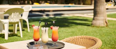 A table with three drinks and a menu, set outdoors with a pool and chairs in the background.