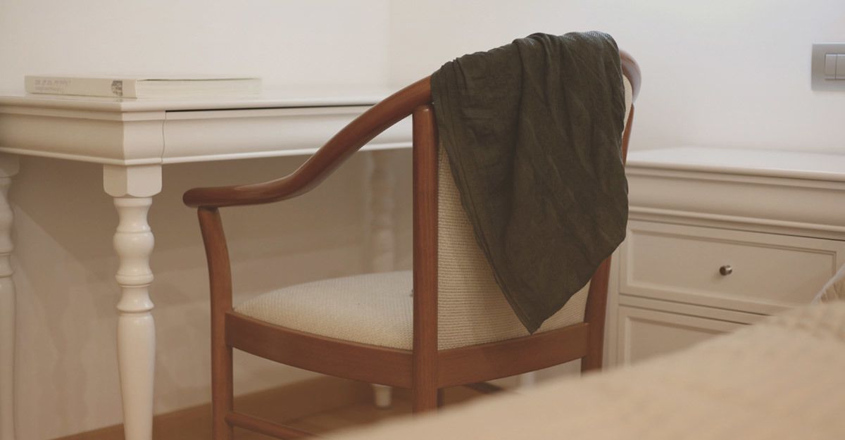 Una silla de madera con un cojín y una manta sobre ella, ubicada frente a un escritorio blanco con un libro encima.