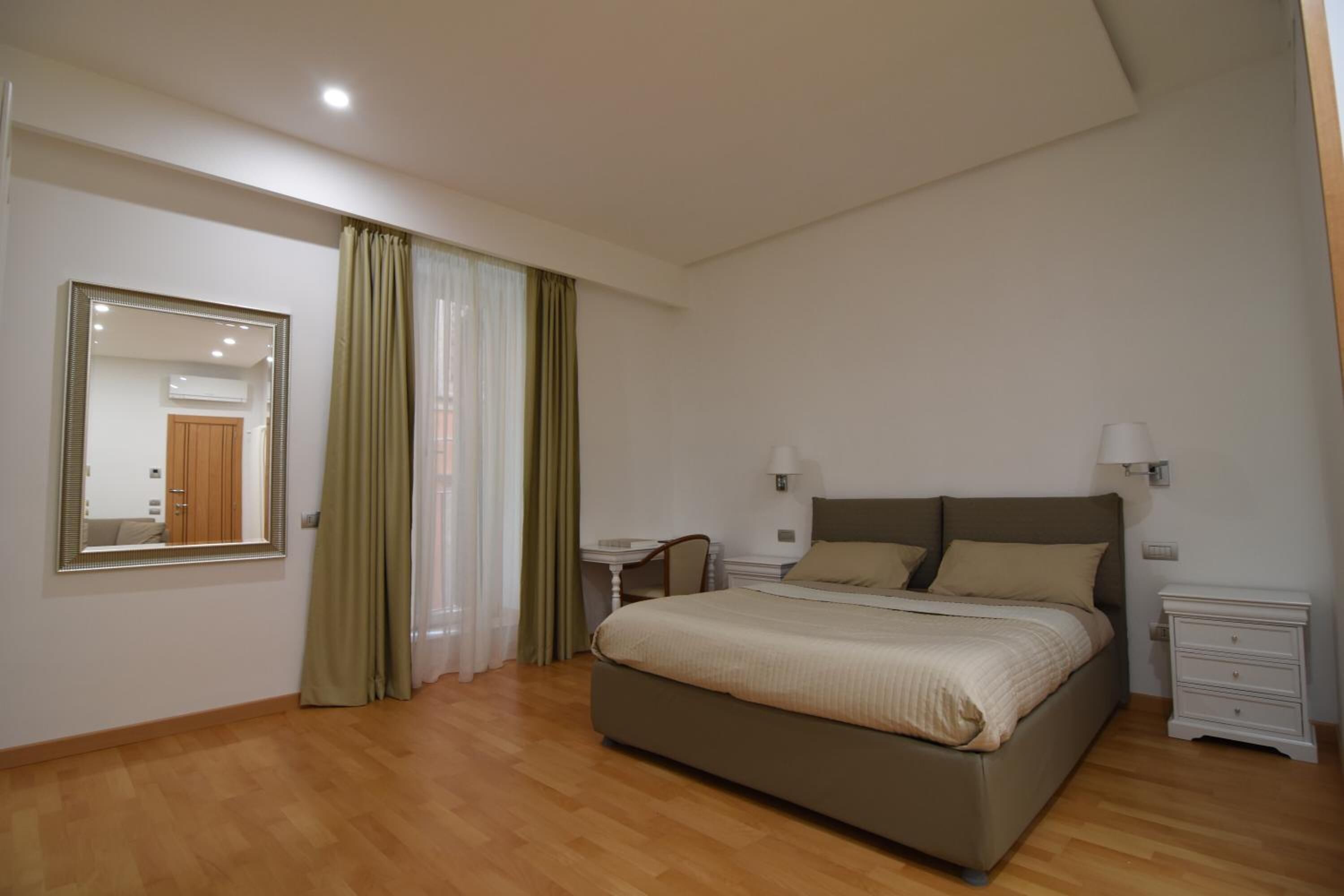 Un dormitori amb un llit gran, una taula de nit, un mirall i una finestra amb cortines.
